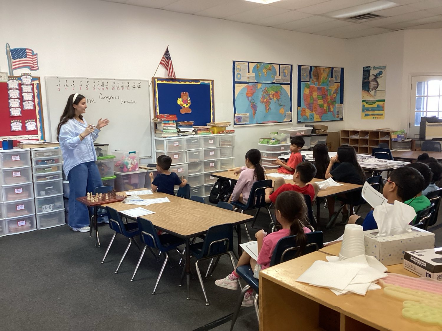 A YCN volunteer stands at a whiteboard reading "HOR, Congress, Senate" and speaks to a classroom of elementary students seated at tables.
