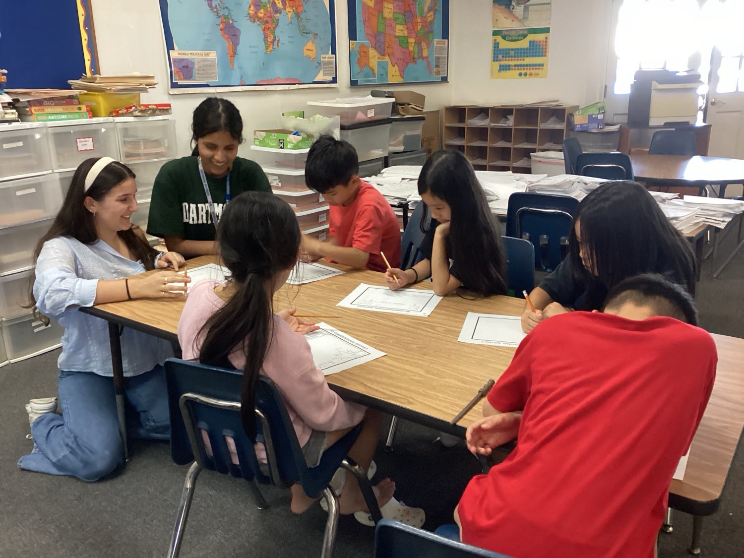 Two YCN volunteers sit at a table with six students who are drawing on worksheets titled "My Congressional District."